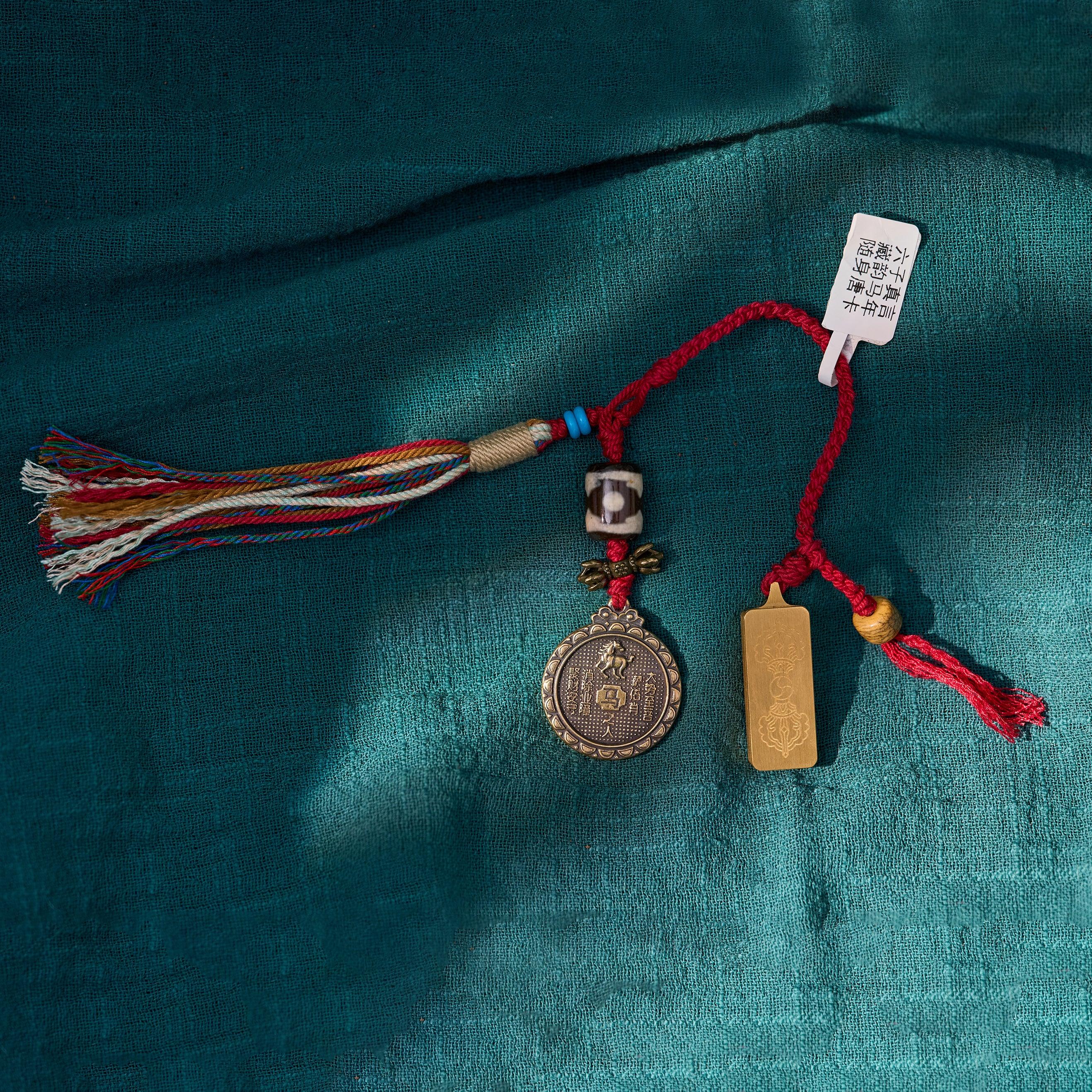 Tibetan-style amulet with red cord, Dzi bead, coin charm, gold tag, and multicolor tassel on teal cloth