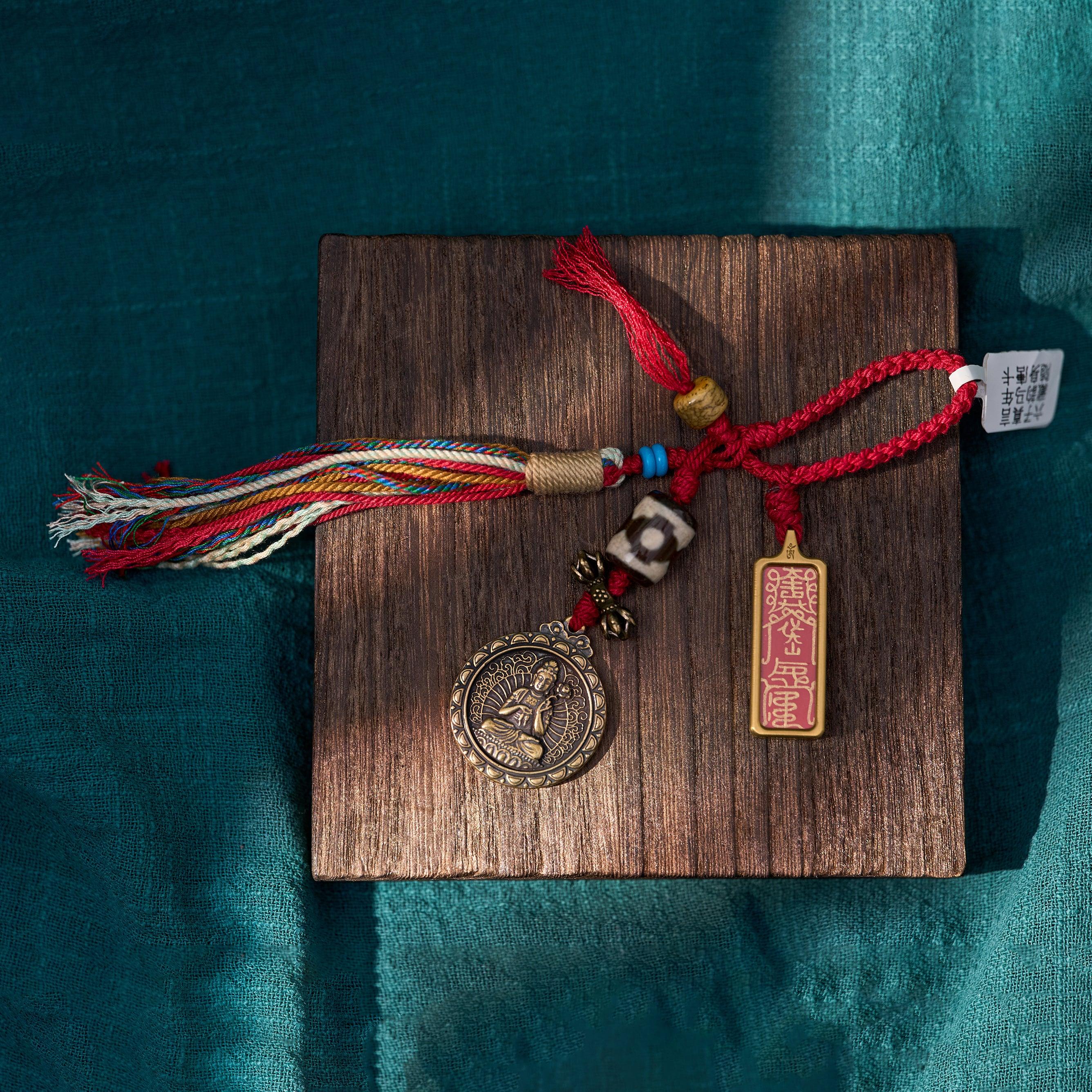Tibetan-inspired handmade charm with red cord, beads, multicolor tassel, and engraved pendant on wooden board