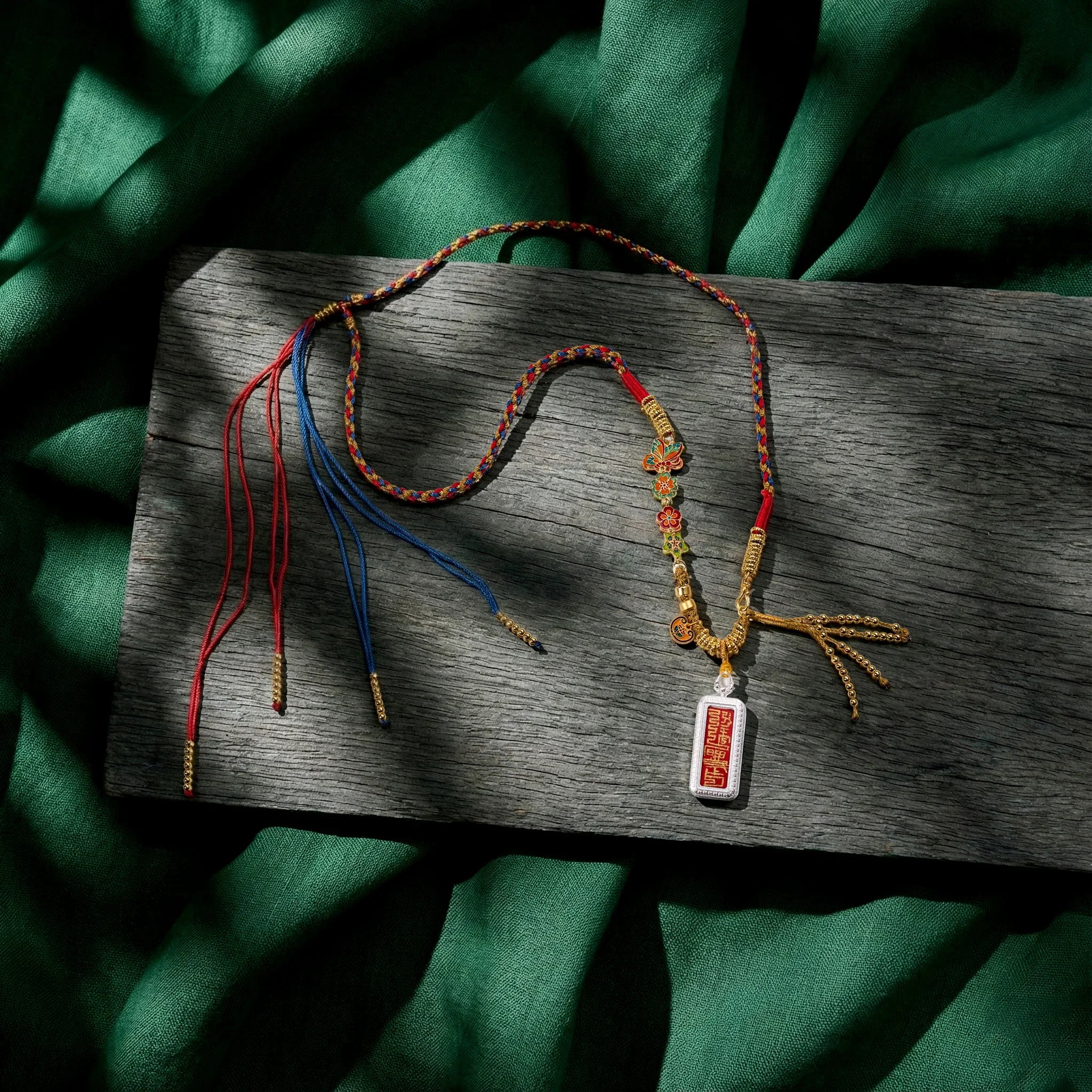 Handcrafted Tibetan necklace with red and blue woven cord, gold accents, and engraved pendant on wood.